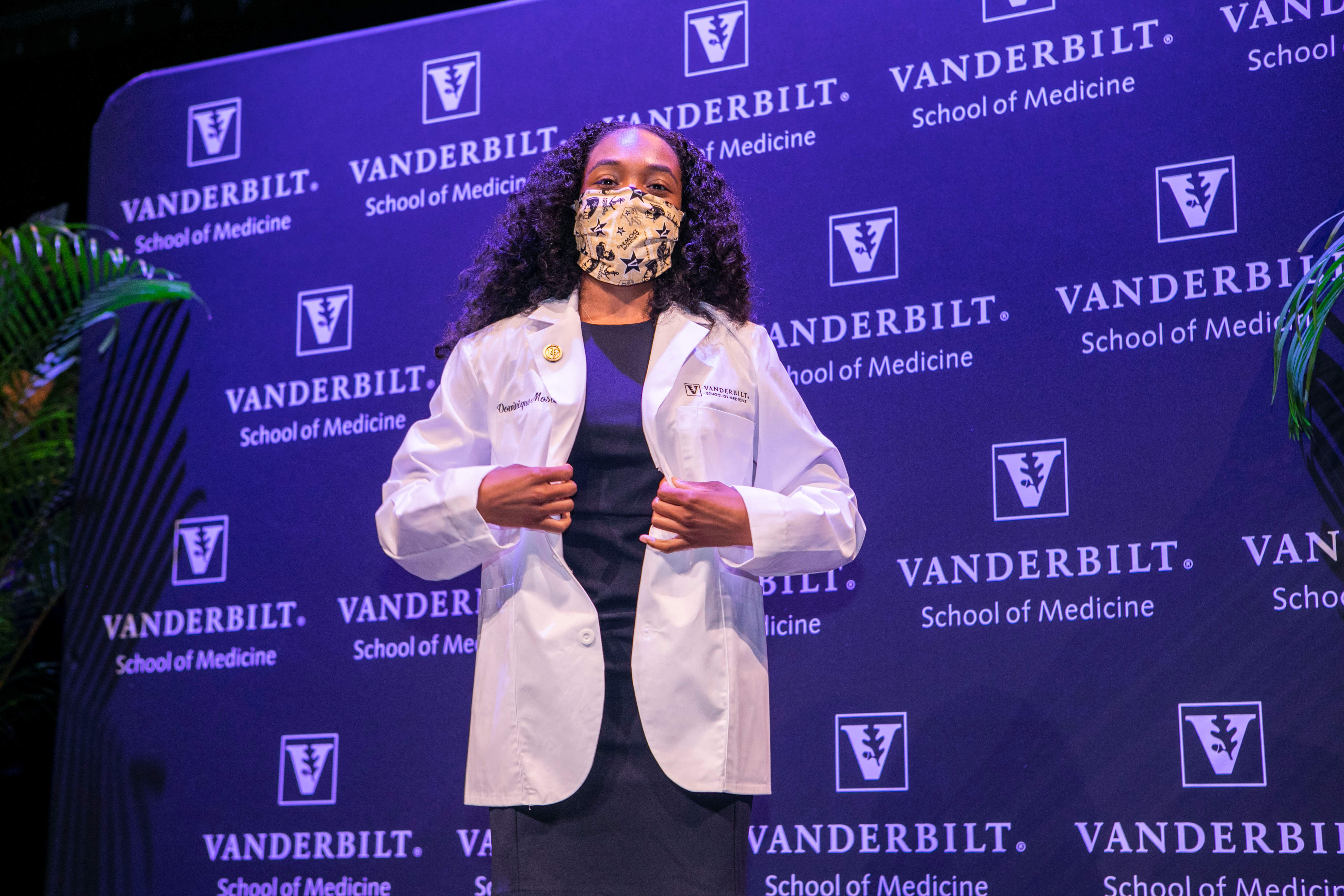 A woman in a mask and white coat stands in front of a medical school backdrop