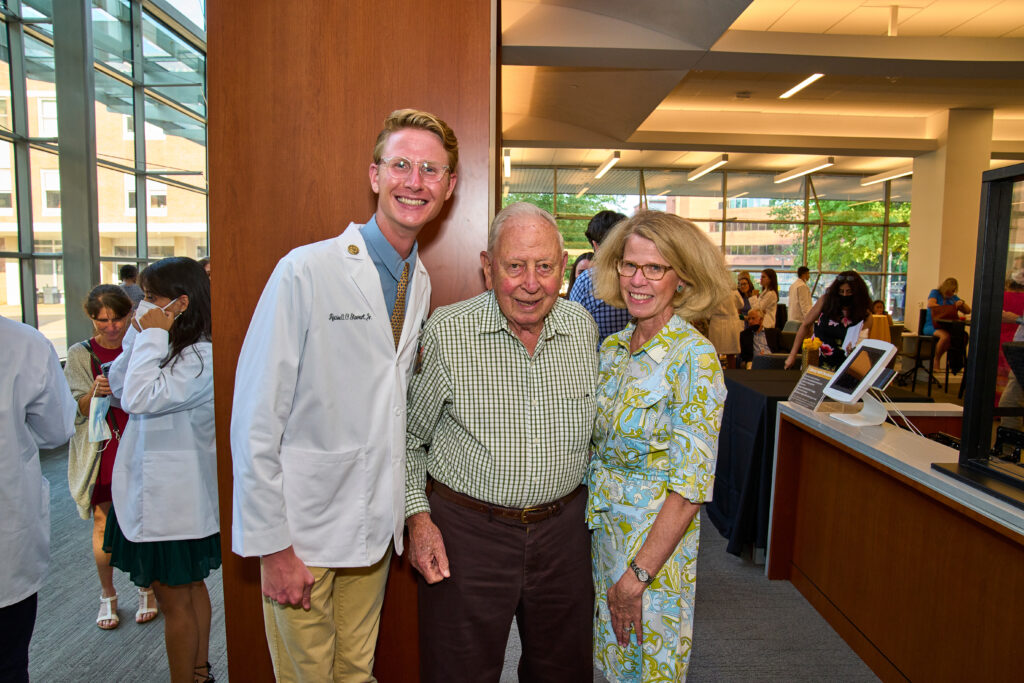 A young man in a short white coat stands with his grandfather and mother in a biomedical library