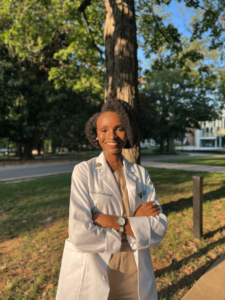 A Black woman in a white coat stands in front of a tree in the early evening sun