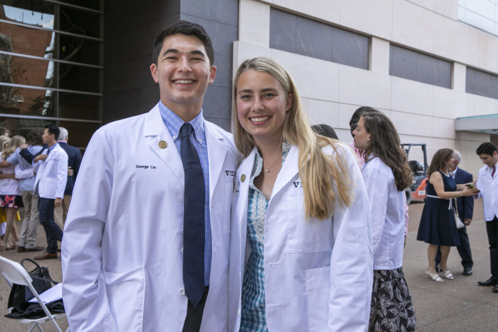 A husband and wife duo in white coats stands outside a medical school