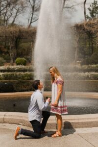 A man drops to one knee and proposes in front of a fountain