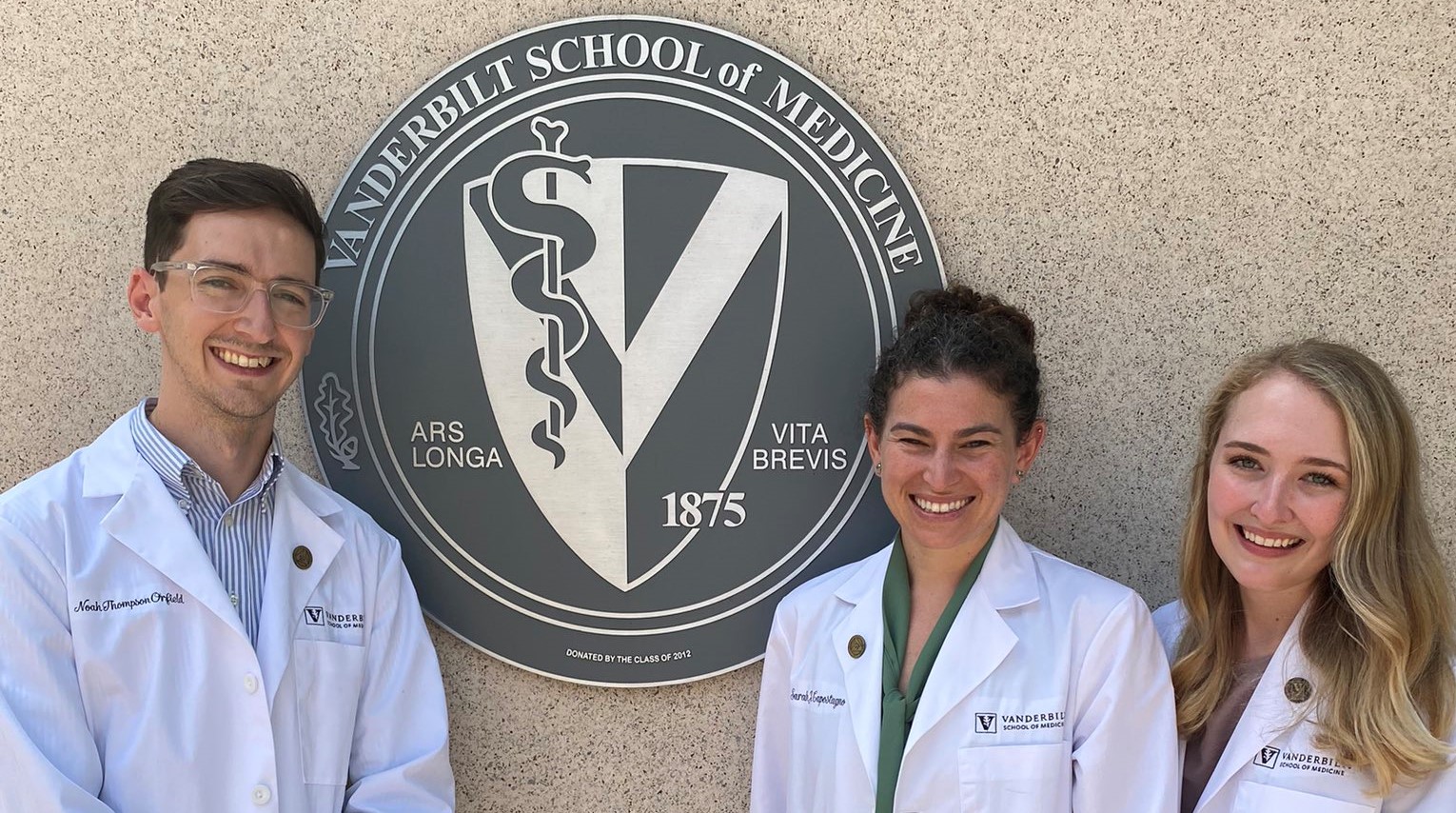 Three medical students in white coats stand next to a medical school seal