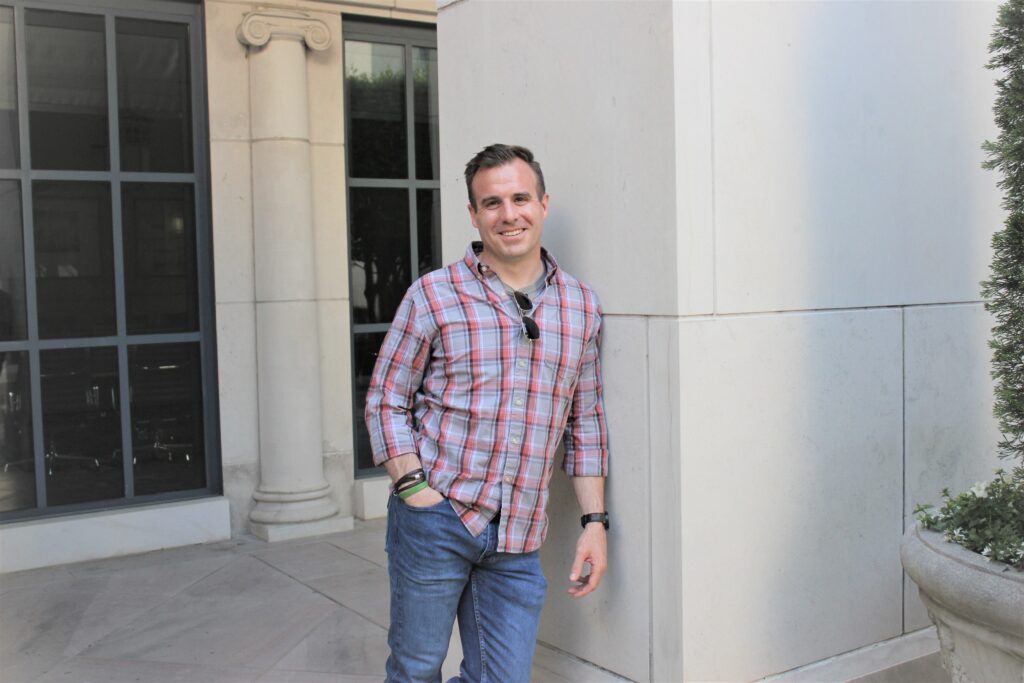 A man in a button down shirt and jeans rests against a wall