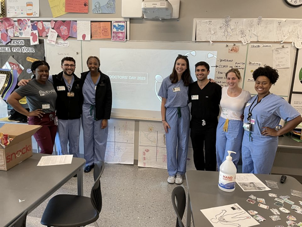  MD student volunteers stand on either side of a classroom screen