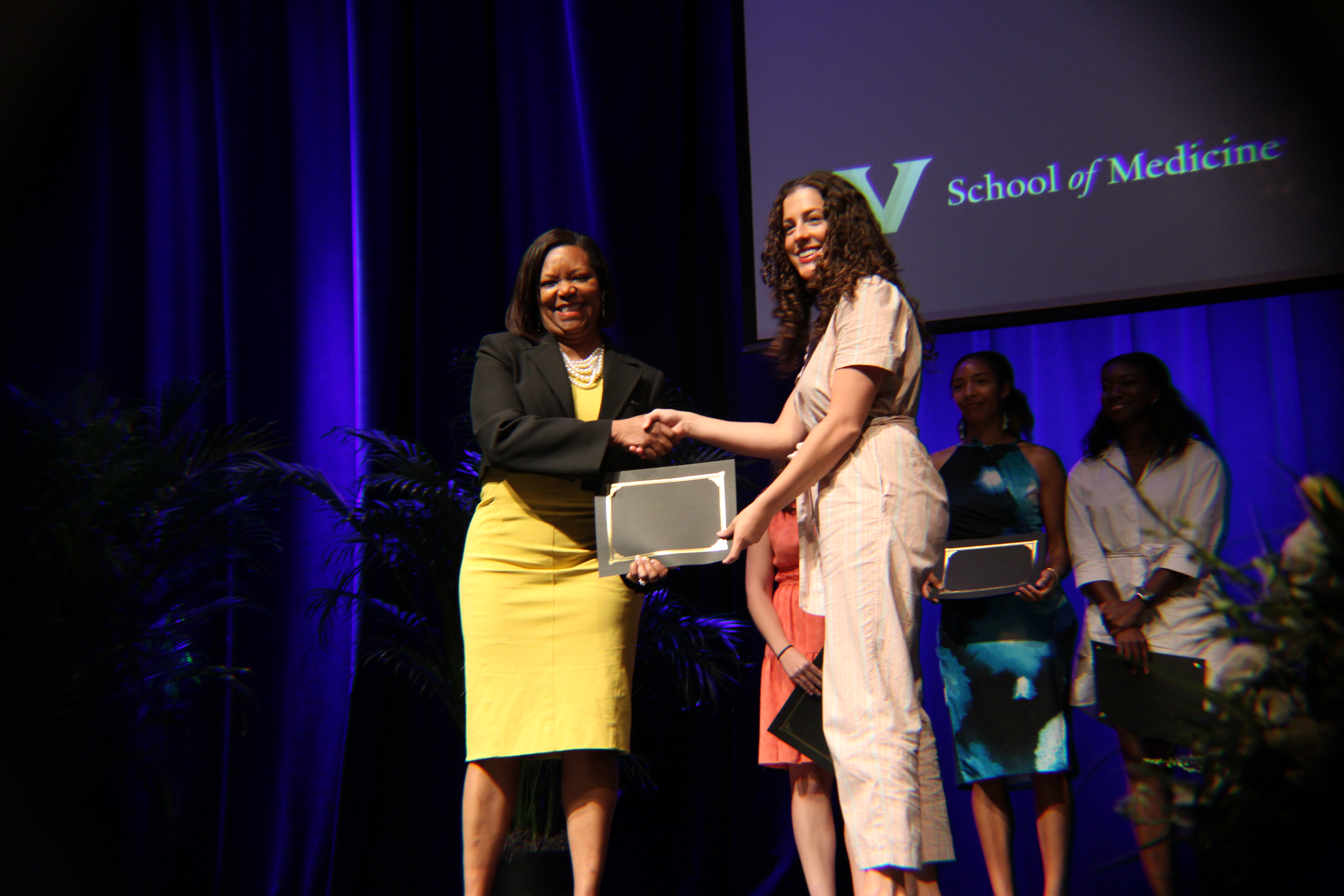A dean in a yellow dress presents an award to an MD student