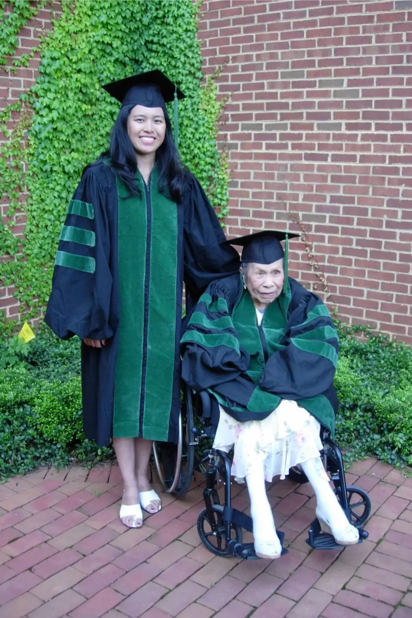 A female medical school graduate poses with her grandmother while both wear academic regalia.