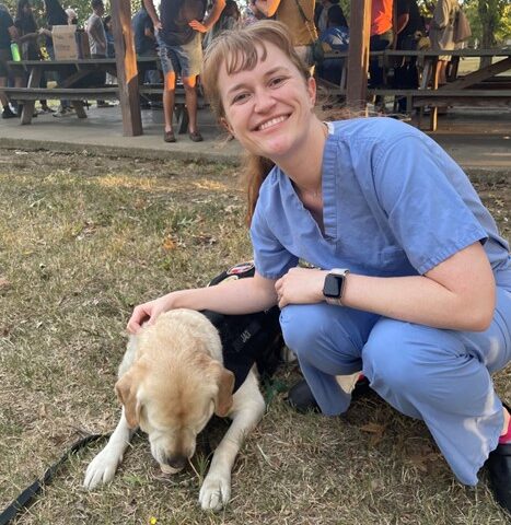 Alison Swartz smiles while kneeling next to and petting a dog.