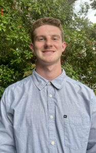 A man in a blue button up stands outside and smiles