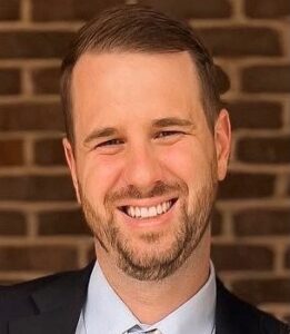 A man in a suit and tie smiles in front of a brick background