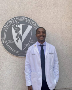 A man in a white coat stands next to a medical school seal outside of a library