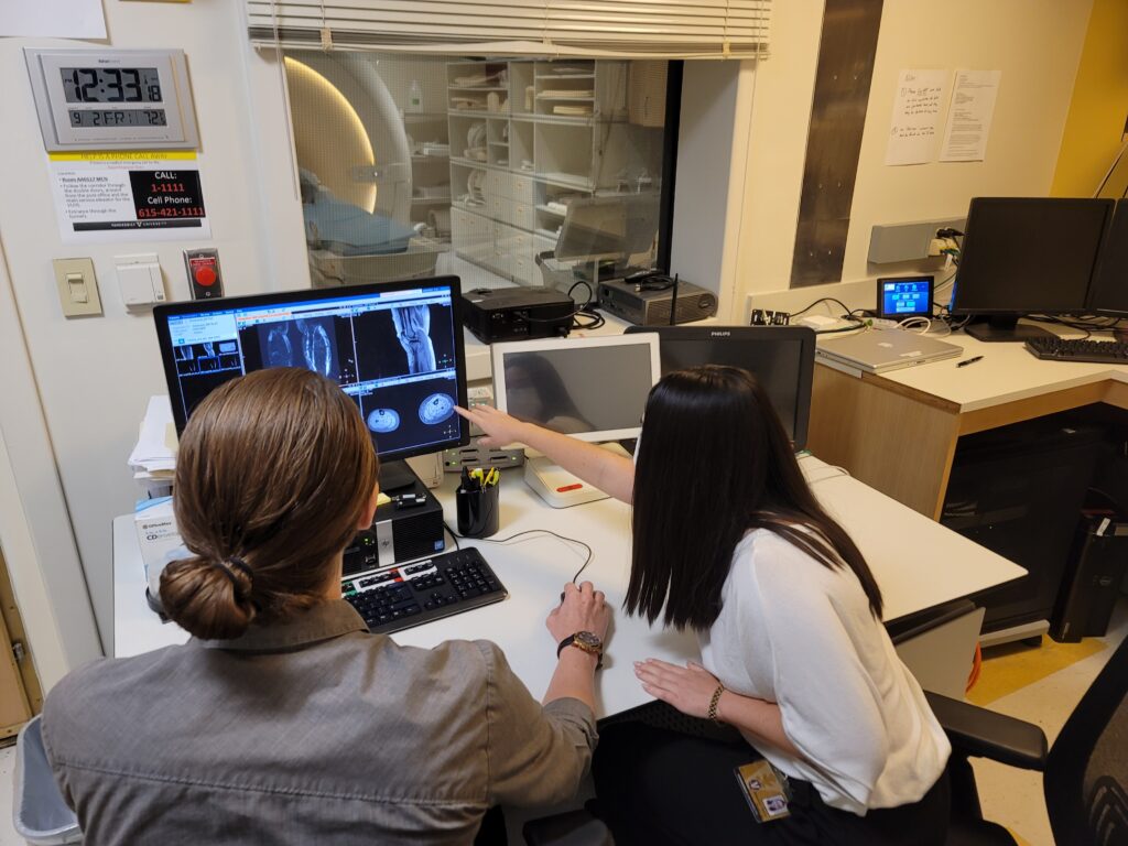 Two women examine scans on a screen while a patient is in an MRI bay