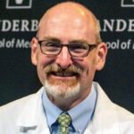 A man in a white coat, green tie, blue shirt, and glasses smiles