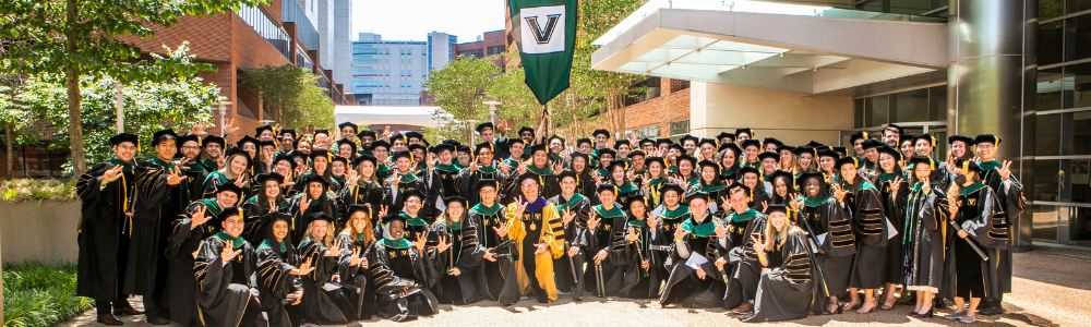 A group of graduates in doctoral regalia pose with the dean (also in doctoral regalia) outside on the medical plaza