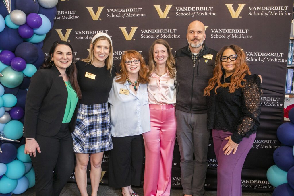 Five women and one man stand in front of a step and repeat banner