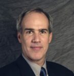 a man in a suit and tie sits in front of a gray backdrop