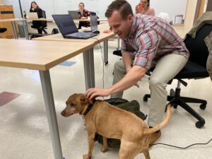 A man pets a dog in a classroom while others sit at a large table
