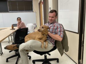 A dog sits on a man's lap as a classmate looks on