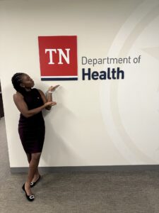 Anjola Ajayi points at a sign on a wall that reads "TN Department of Health"
