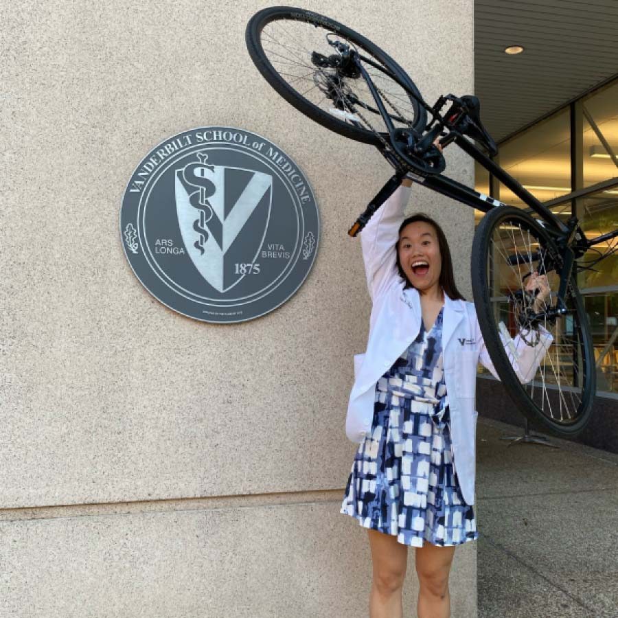 A female medical student in a white coat holds a bicycle above her head.