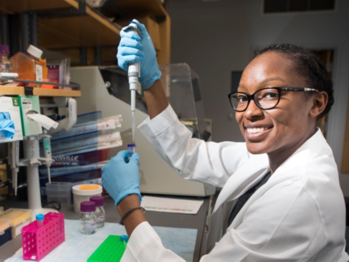 A smiling female student researcher in a lab coat and gloves pipettes a liquid in a VUSM lab.